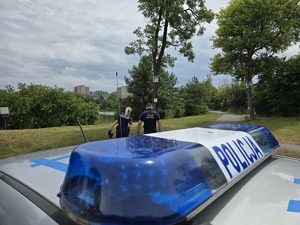 zdjęcie przedstawia zbliżenie na radiowóz a w tle policjantka i strażnik miejski