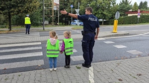 zdjęcie przedstawia: policjanta i dzieci z przedszkola uczące się przechodzić przez jezdnię
