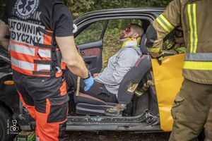zdjęcie przedstawia poszkodowanego we wraku samochodu, obok strażaka i medyka