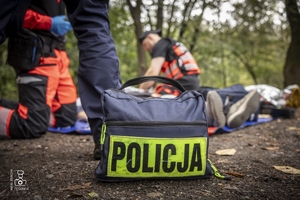 zdjęcie przedstawia zbliżenie na apteczkę a w tle poszkodowany i medycy udzielających mu pomocy