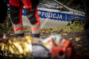 zdjęcie przedstawia zbliżenie na radiowóz a na rozmazanym pierwszym planie medycy i poszkodowany
