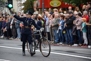 Na fotografii widać mężczyznę, który idzie ulicą i prowadzi rower. Ubrany jest w stary policyjny mundur. Po obydwu stronach ulicy znajduje się tłum ludzi.