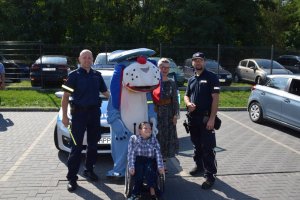 Na fotografii wdać stojących przed radiowozem policjantów, policyjną maskotkę Sznupka i Panią Dyrektor Ośrodka w Ziemięcicach. Przed nimi na wózku inwalidzkim siedzi niepełnosprawny chłopiec.
