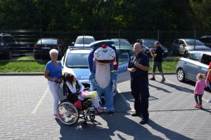 Na fotografii wdać stojących przed radiowozem policjanta, policyjną maskotkę Sznupka oraz kobietę i dziewczynkę siedzącą na wózku inwalidzkim.