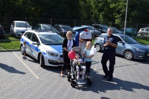 Na fotografii wdać stojących przed radiowozem policjanta, policyjną maskotkę Sznupka oraz kobietę i dziewczynki, z których jedna siedzi na wózku inwalidzkim.