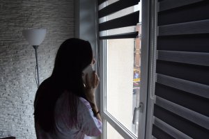 Przy oknie stoi młoda dziewczyna, która przy uchu trzyma telefon komórkowy.