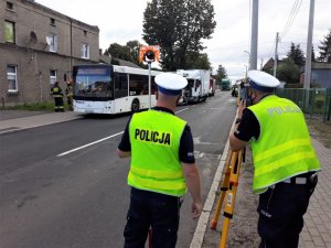 zdjęcie przedstawia dwóch policjantów stojących obok urządzenia służącego do wykonywania pomiarów. W tle widać uszkodzony autobus oraz ciężarówkę.