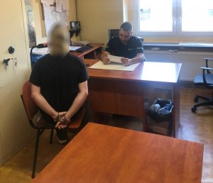 Na zdjęciu widać pomieszczenie znajdujące się w Komisariacie Policji. Przy biurku siedzi policjant, a po drugiej stronie zatrzymany mężczyzna, który na rękach trzymanych z tyłu ma zapięte kajdanki
