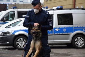 Na zdjęciu widać policjanta, który trzyma trzyma psa na smyczy. W tle zdjęcia widać policyjne radiowozy.