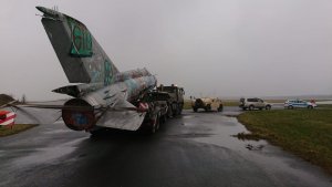Na zdjęciu widać samolot wojskowy MIG-21 wieziony na przyczepie ciągniętej przez samochód ciężarowy. Przed ciężarówką jadą inne pojazdy, na których czele jedzie policyjny radiowóz.