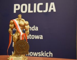 Na zdjęciu widać złoty medal i statuetkę strongmena. W tle znajduje się baner z napisem Policja