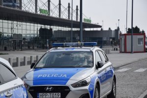 Na zdjęciu widać policjanta stojącego obok radiowozów znajdujących się na parkingu lotniska.