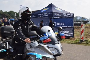 Na zdjęciu widać policjanta na motocyklu. W tle widać policyjny namiot profilaktyczny oraz będącego pod nim policjanta.