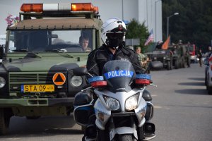 Na zdjęciu widać policjanta na motocyklu, za którym ustawione są jeden za drugim wojskowe samochody.
