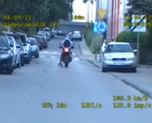 Na zdjęciu widać jadącego ulicą motocyklistę. Na poboczu jezdni widać zaparkowane radiowozy.