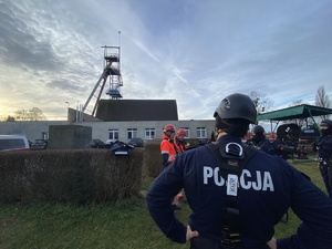 na zdjęciu policjant w mundurze i uprzęży do pracy na wysokości obserwuje znajdującą się w oddali wieżę szybową kopalni