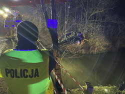 na zdjęciu policjant w odblaskowej kamizelce z napisem policja, stojąc na moście, obserwuje akcję wyciągania samochodu z rzeki