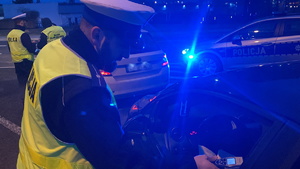 na zdjęciu policjant w trakcie badania trzeźwości kierującego, w tle radiowóz emitujący niebieskie światła błyskowe