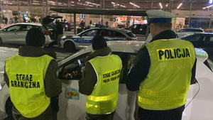 na zdjęciu od lewej funkcjonariusz Straży Granicznej, Służby Celno-Skarbowej i Policji w trakcie kontroli białego pojazdu, w tle widoczny policyjny radiowóz i fragment terminala lotniska