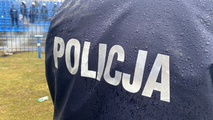na zdjęciu fragment niebieskiego munduru z napisem policja, w oddali część stadionowego sektora