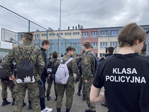na zdjęciu widoczny uczeń w czarnej koszulce z napisem klasa mundurowa, stojący tyłem do obiektywu, przed nim inne osoby, a w oddali budynek szkoły, na którego dachu stoją policjanci