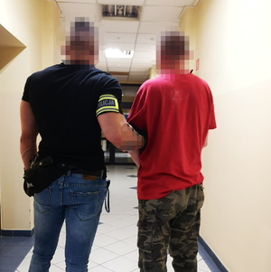 na zdjęciu policjant z opaską na ramieniu prowadzi korytarzem komendy zatrzymanego mężczyznę w czerwonej koszulce