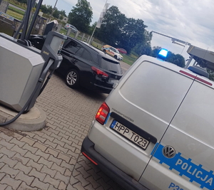 na zdjęciu policyjny radiowóz z włączonymi niebieskimi sygnałami, stojący tyłem obok zaparkowanego czarnego pojazdu, za nimi inny kolorowy pojazd i drzewa
