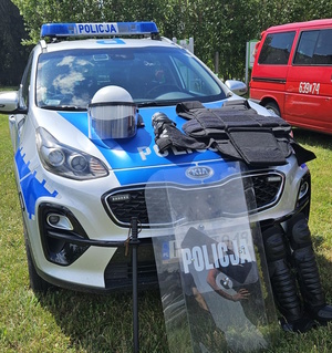na zdjęciu policyjna tarcza, kask i ochraniacze oparte o policyjny radiowóz, obok fragment wozu strażackiego