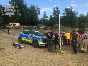 na zdjęciu na plaży przy wodzie radiowóz, policjanci, ratownicy i strażacy przy parawanie