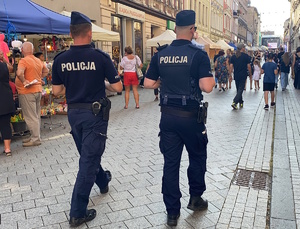 na zdjęciu policjanci w trakcie patrolu pieszego na jednej z ulic miasta, gdzie widać osoby i namioty, w oddali scena na rynku