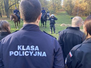 na zdjęciu osoba w mundurze z napisem na plecach klasa policyjna, przed nią policjanci na koniach