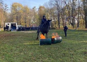 na zdjęciu policyjny jeździec pokonuje na koniu palącą się przeszkodę na terenie parku, w oddali zgormadzona młodzież