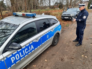 na zdjęciu policjant przy radiowozie, w rękach trzyma urządzenie sterujące dronem