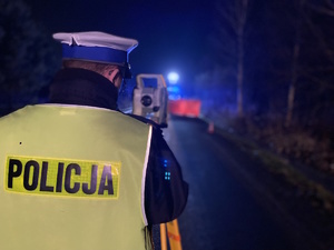 na zdjęciu policjant przy urządzeniu na jezdni po zmroku, przed nim miejsce zdarzenia i widoczny czerwony parawan