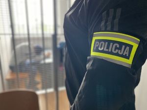 na zdjęciu fragment policjanta po cywilu z opaską na ramieniu z napisem policja, w oddali w celi zatrzymany