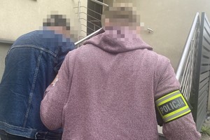 na zdjęciu policjant po cywilu z opaską na ramieniu, prowadzi do budynku komendy zatrzymanego