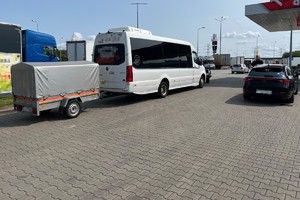na zdjęciu biały bus z przyczepą na terenie stacji benzynowej, obok nieoznakowany radiowóz