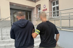 na zdjęciu policjant z opaską na ramieniu z napisem policja prowadzi zatrzymanego do budynku prokuratury