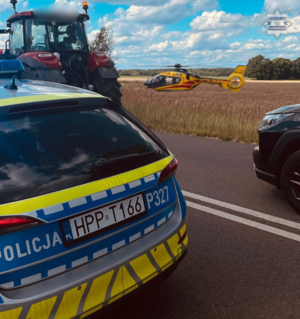 na zdjęciu radiowóz na jezdni, obok w polach śmigłowiec lotniczego pogotowia ratunkowego
