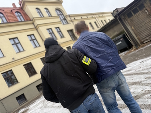 na zdjęciu policjant po cywilu z opaską prowadzi zatrzymanego