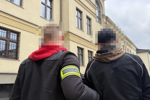 na zdjęciu policjant z opaską na ramieniu z napisem policja, prowadzący zatrzymanego mężczyznę