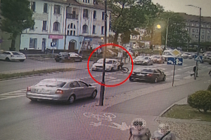 na zdjęciu fragment drogi, na której są osoby i pojazdy, a w czerwonym kółku zaznaczona została rowerzystka, przy pojeździe po uderzeniu w nie