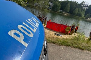 na zdjęciu fragment policyjnego radiowozu, w oddali na plaży strażacki parawan i ratownicy wokół niego