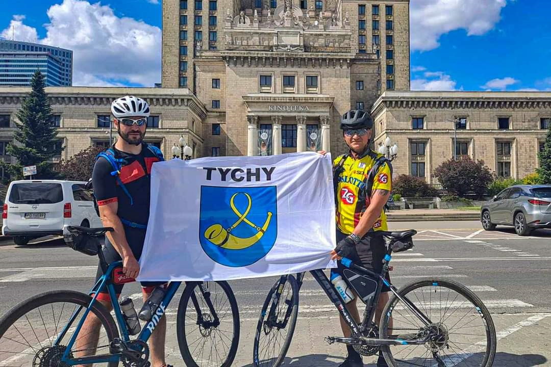 Dwaj mężczyźni przy rowerach trzymają flagę Tychów na tle Pałacu Kultury.