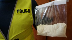 Obrazek przestawia policjanta w kamizelce odblaskowej z napisem &quot;Policja&quot; trzymającego worek z białym proszkiem-amfetaminą
