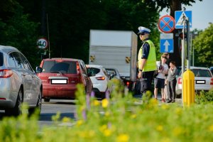 Obrazek przedstawia policjanta stojącego wzdłuż drogi, na której jadą samochody, w tle widoczni inni ludzie