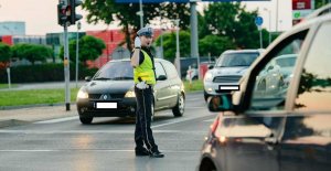 Na obrazku widoczny policjant wydziału ruchu drogowego, w białych rękawiczkach oraz z gwizdkiem, stoi na skrzyżowaniu i kieruje ruchem
