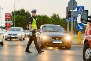 Na obrazku widoczny policjant wydziału ruchu drogowego, w białych rękawiczkach oraz z gwizdkiem, stoi na skrzyżowaniu i kieruje ruchem