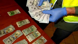 Nieumundurowany policjant w odblaskowej kamizelce i rękawiczkach trzyma w plik dolarów. W tle widoczne na burku pieniądze.