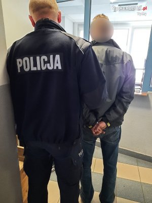 z lewej strony stoi umundurowany policjant z prawej strony stoi zatrzymany mężczyzna, który ma założone kajdanki na ręce trzymane z tyłu